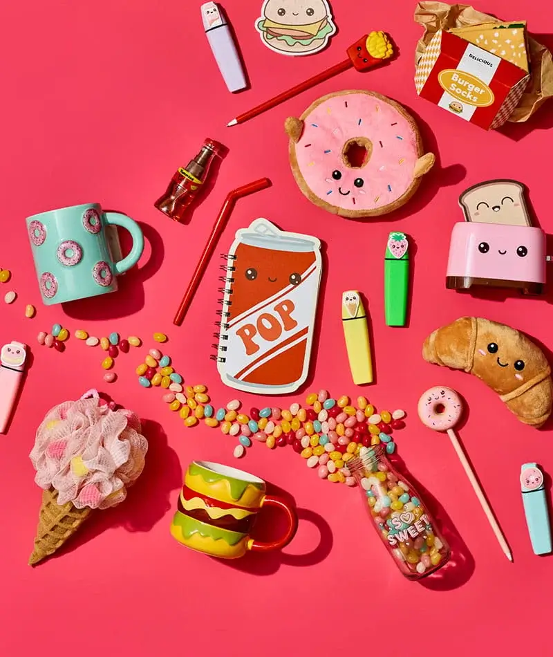 A colorful flat lay on a pink background featuring playful food-themed items like plush donuts, novelty mugs, stationery, sweets, and cute kitchen accessories.
